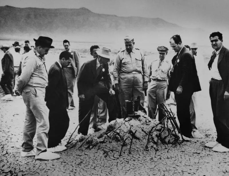 Robert Oppenheimer and General Leslie Groves and others examine the warped wreckage that is all that remains of a hundred-foot tower and shack that held the first nuclear weapon.