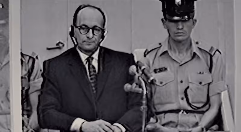 Adolf Eichmann sits expressionless inside a glass booth during his 1961 trial in Jerusalem, flanked by Israeli guards, as depicted in the 2012 biographical film Hannah Arendt.