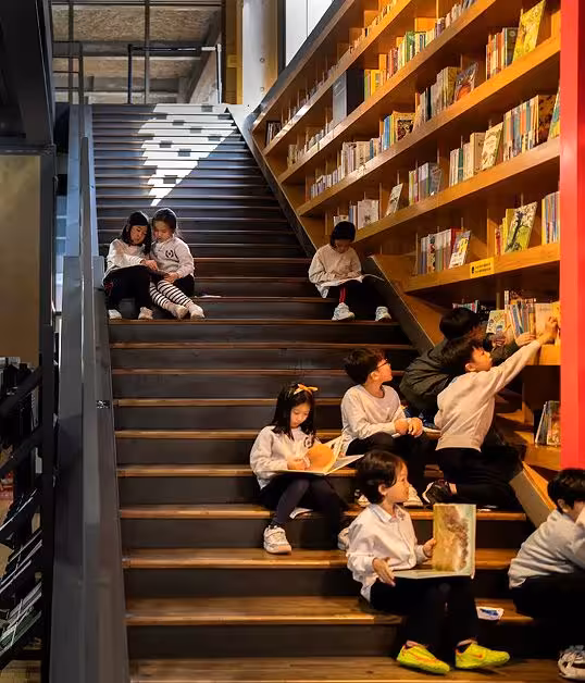 Children reading and exploring books on the wooden staircase inside the Forest of Wisdom, the vast public library and central attraction of Paju Book City in South Korea — a space where learning, curiosity, and literature come alive through immersive design